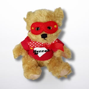 May include: A plush teddy bear wearing a red superhero mask and shirt. The shirt has white polka dots and the name "Hannah" in a diamond shape. The bear has tan fur and a black nose.