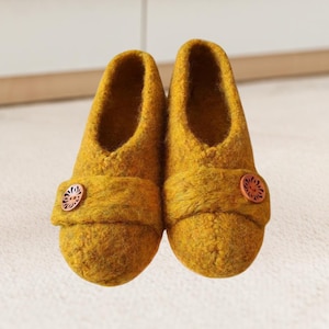 Puede incluir: Un par de zapatillas de lana de color amarillo mostaza con un detalle de botón marrón. Las zapatillas tienen una punta redonda simple y una suela plana.