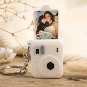 May include: A white camera-shaped keyring with a silver ring. The keyring features a small photo of two people. The camera has a black lens and a silver button. The background is a wooden surface with dried flowers.