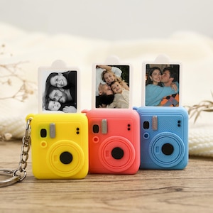 May include: Three miniature camera keyrings in yellow, coral, and blue. Each holds a small photograph. The keyrings are displayed on a wooden surface, with a soft, white background.
