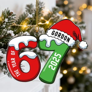 Puede incluir: Adorno navideño con un número 6 rojo y un número 7 verde, ambos con contorno blanco y cubiertos de nieve. El adorno tiene el texto "THE YEAR OF" y "2025". Un gorro de Papá Noel con el nombre "GORDON" está en la parte superior.