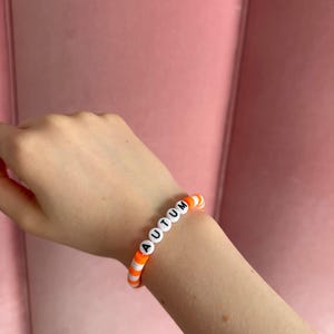 Pu&ograve; includere: Un braccialetto di perline bianche e arancioni con la parola "AUTUMN" scritta in lettere nere.