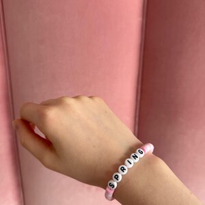 Pu&ograve; includere: Un braccialetto di perline rosa e bianche con la parola "SPRING" scritta in lettere nere.