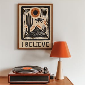 Peut inclure: Une impression encadrée avec un motif de paysage désertique représentant un soleil, des cactus et une soucoupe volante. L'impression porte le texte "I BELIEVE" en bas. Un tourne-disque vintage et une lampe rouge sont également présents sur l'image.