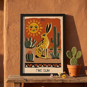 May include: Framed art print featuring a yellow and black spotted jackrabbit, cacti, and a smiling sun against an orange background. The text "THE SUN" is at the bottom. The artwork is in a black frame.