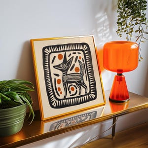 May include: A gold framed print with a black and white illustration of a stylized dog with orange accents. The print is on a wooden shelf with an orange glass lamp.
