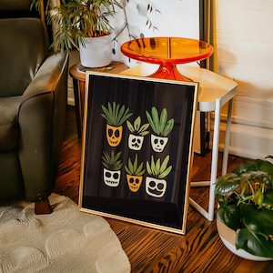 Puede incluir: Una impresión enmarcada con fondo negro y marco dorado. La impresión presenta una ilustración caprichosa de seis plantas en maceta con caras de calavera sonrientes. Las plantas son verdes y naranjas.