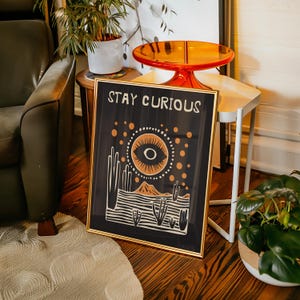 May include: A framed print with a gold frame and a black background. The print features a stylized eye with a mountain range and cacti in the background. The text "STAY CURIOUS" is at the top of the print.