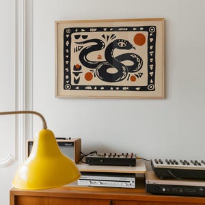 Op de afbeelding: Een ingelijste print van een zwarte slang met geometrische patronen op een crèmekleurige achtergrond. De slang is opgerold met zijn kop omhoog en heeft een groot oog. De print is omgeven door een houten lijst.