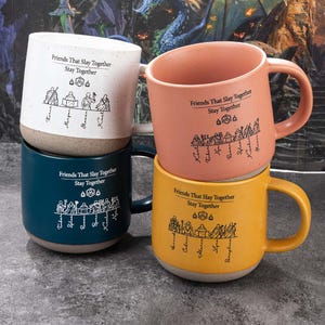May include: Four ceramic mugs in white, dark blue, peach, and yellow, each featuring a speckled base and a black graphic print. The design includes the text "Friends That Slay Together Stay Together," a twenty-sided die icon, and a line of fantasy characters.