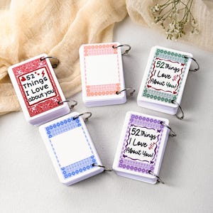 Op de afbeelding: Vijf kleine, witte notitieblokken met decoratieve randen en metalen ringen. Elk notitieblok heeft een andere gekleurde rand: rood, roze, blauw, groen en paars. Op het rode notitieblok staat "52 dingen die ik leuk vind aan jou."