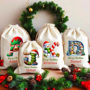 May include: Five off-white drawstring Christmas gift bags with festive designs. Each bag features a unique holiday graphic, including an elf, a Christmas tree, a snowman, and a letter. The bags are decorated with red text and a green wreath.