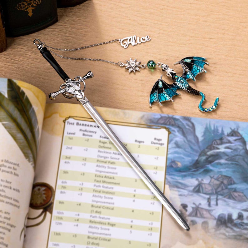 May include: A silver sword-shaped bookmark with a black handle rests on an open book. A dragon-shaped charm with teal wings and a nameplate that says "Alice" are also present. The bookmark is designed for fantasy book lovers.