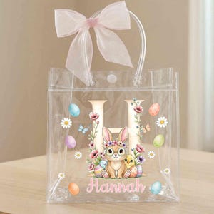 Puede incluir: Un bolso de mano de plástico transparente con una letra H grande, un conejo con corona de flores, un pollito y huevos de Pascua coloridos. El bolso está decorado con margaritas y mariposas, incluye el nombre Hannah y un lazo rosa claro.