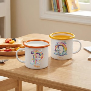 Pode incluir: Duas canecas de esmalte branco com desenhos de arco-íris e nomes personalizados. Uma caneca tem a letra "P" com um arco-íris e o nome "Poppy". A outra caneca tem um desenho de arco-íris e o nome "Oliver". Ambas as canecas têm uma borda colorida.