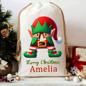 May include: A white drawstring Christmas gift bag featuring a colorful elf design in the shape of the letter "A". The elf wears a red hat and green outfit. The text "Merry Christmas Amelia" is printed in green and red.