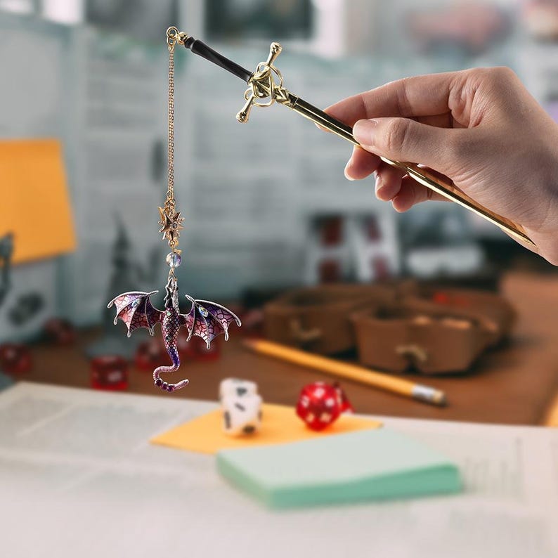 May include: A fantasy-themed bookmark featuring a miniature dragon with purple and red wings, suspended from a gold-colored sword. The sword is held by a hand, with dice and other game accessories in the background.