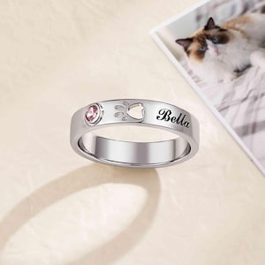 Peut inclure: Bague argentée ornée d'une pierre rose, d'une empreinte de patte et du nom "Bella". La bague est probablement un accessoire sur le thème des animaux de compagnie, peut-être un hommage à un chat.