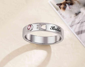 Puede incluir: Un anillo plateado con una gema rosa, una huella de pata y el nombre "Bella". El anillo es probablemente un accesorio temático para mascotas, posiblemente un recuerdo o un homenaje a un gato.