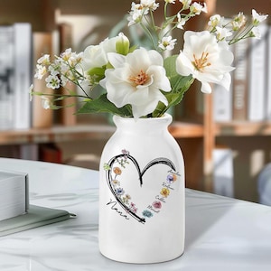 Puede incluir: Un jarrón de cerámica blanco con un diseño floral en forma de corazón y la palabra "Nana". El jarrón contiene flores blancas con hojas verdes. El diseño del corazón incluye flores coloridas y nombres.