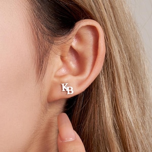 Può includere: Orecchini a lobo in argento con le iniziali "KB" su un orecchio. Gli orecchini sono piccoli e minimalisti, riflettendo uno stile di gioielleria moderno. L'orecchio è posizionato su uno sfondo sfocato.