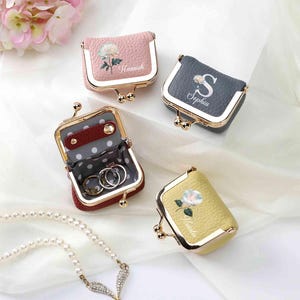May include: Four small, personalized ring boxes in pink, gray, and yellow, with gold-colored metal frames. Each box features a floral design and a name. One box is open, revealing rings inside. A pearl necklace is in the foreground.