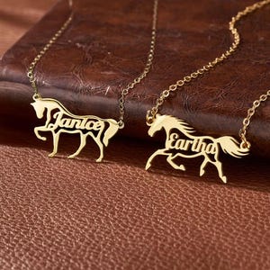 May include: Two gold-toned necklaces featuring a horse-shaped pendant. Each pendant is cut out in the silhouette of a horse with the name Janice or Eartha integrated into the body of the horse. The necklaces have thin gold-toned chains.