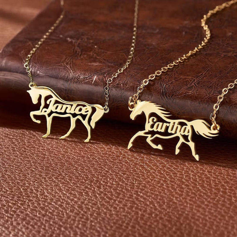 May include: Two gold-toned necklaces featuring a horse-shaped pendant. Each pendant is cut out in the silhouette of a horse with the name Janice or Eartha integrated into the body of the horse. The necklaces have thin gold-toned chains.