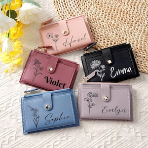 Könnte beinhalten: Fünf personalisierte Geldbörsen in den Farben Rosa, Burgunderrot, Schwarz, Blau und Lavendel. Jede Geldbörse hat ein Blumendesign und einen Namen: Hazel, Violet, Emma, Sophia und Evelyn. Die Geldbörsen haben einen Druckknopfverschluss und ein Reißverschlussfach.