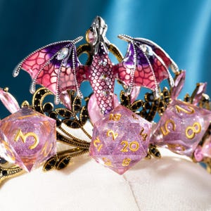 Könnte beinhalten: Eine Fantasy-Krone mit einem Drachen und Würfeln. Die Krone hat einen silbernen Drachen mit rosa und lila Flügeln und schwarzen und goldenen Details. Die Würfel sind transparent lila mit goldenen Zahlen, ideal für Tabletop-Spiele.