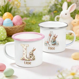 May include: Two white enamel mugs with Easter designs. One has a pink rim, the letter "L", a bunny holding an egg, and the name "Lillie". The other features the letter "A", a bunny, and the name "Alice".
