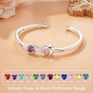 Puede incluir: Un brazalete infinito plateado con dos piedras preciosas en forma de corazón, una púrpura y otra rosa. Los nombres "Kaitlyn" y "Evelyn" están grabados en el brazalete. Se muestra una selección de colores de piedras de nacimiento debajo.