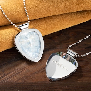 Könnte beinhalten: Zwei silberne Gitarrenplektrum-Halsketten. Eine hat einen perlmuttartigen Einsatz, die andere den eingravierten Text "I Pick You". Beide sind an Kugelkettchen. Der Hintergrund ist braunes Leder und Holz.