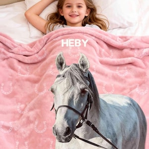 Könnte beinhalten: Eine rosa Decke mit einem Hufeisenmuster und dem Namen "HEBY" in weißen Buchstaben. Die Decke zeigt ein detailliertes Bild des Kopfes eines weißen Pferdes mit Zaumzeug. Ein junges Mädchen liegt unter der Decke auf einem Bett.
