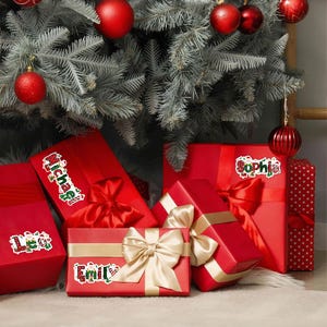 May include: Red wrapped Christmas gifts with gold and red bows are arranged under a decorated Christmas tree. Each gift has a name sticker: Michael, Emily, Sophia, and Leo. Red ornaments hang from the tree.