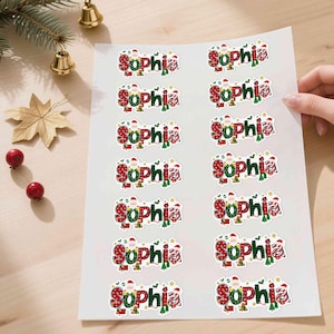 May include: Sheet of stickers with the name "Sophia" in a festive Christmas design. The stickers feature red and green plaid patterns, Santa Claus illustrations, and gold stars. The stickers are on a white sheet.