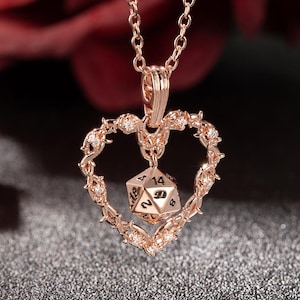 May include: Rose gold necklace featuring a heart-shaped pendant with a dice charm. The heart is adorned with small crystals and a thorny design. The dice charm has visible numbers. The necklace is suspended from a delicate chain.