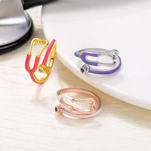 May include: Three gold rings with a stethoscope design. The rings are in pink, purple, and white enamel.