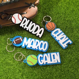 Pode incluir: Uma coleção de chaveiros personalizados com temas esportivos. Os chaveiros incluem uma bola de beisebol, basquete, futebol americano e softball, cada um combinado com um nome em letras brancas. Em uma superfície verde.