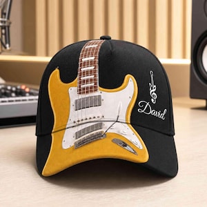 Könnte beinhalten: Eine schwarze Baseballkappe mit einer Grafik einer gelben E-Gitarre mit weißem Schlagbrett und einem braunen Griffbrett, das sich über die Kappe erstreckt. Der Name David ist in weißer Schreibschrift neben einem Notenschlüssel-Gitarrensymbol gedruckt.