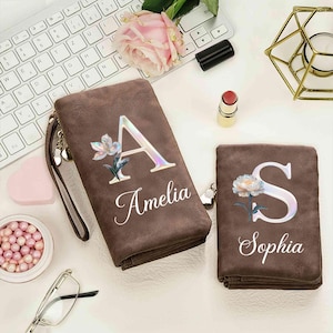 Puede incluir: Dos carteras marrones con diseños florales e iniciales. Una cartera presenta la letra "A" con el nombre "Amelia", y la otra tiene la letra "S" con el nombre "Sophia". Ambas carteras tienen una correa de muñeca y están sobre una superficie blanca.