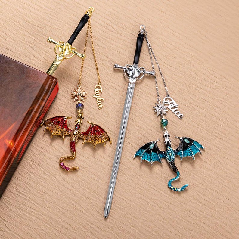 May include: Two sword-shaped bookmarks with dragon charms. One bookmark is gold with a red and gold dragon, the other is silver with a blue and black dragon. Each has a name charm.