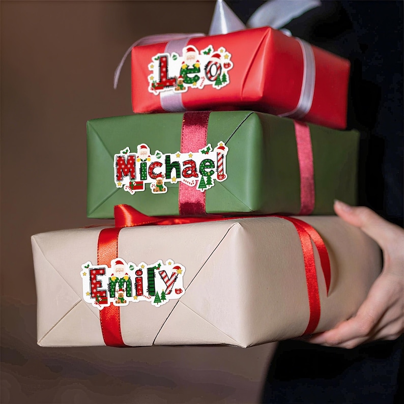 May include: Three wrapped gifts stacked on top of each other. The top gift is red, the middle is green, and the bottom is tan. Each gift has a name sticker with a Christmas theme. The gifts are tied with red ribbon.