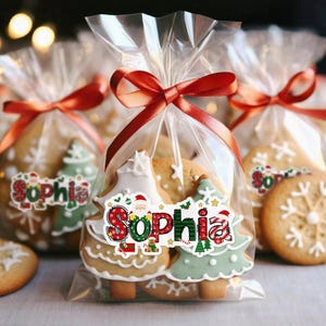 May include: Christmas-themed gingerbread cookies in clear cellophane bags tied with red ribbon. The cookies are decorated with white icing and festive designs, including Christmas trees and snowflakes. Each bag has a sticker with the name "Sophia".