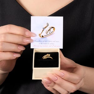 Puede incluir: Un anillo dorado con diseño de ala y pequeñas piedras preciosas azules, presentado en una caja beige. El anillo se muestra con una tarjeta que dice "Forever my love" y una cita romántica. El anillo es una joya.