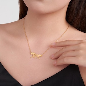 May include: A gold-toned necklace featuring a delicate chain and a pendant shaped like a horse in motion. The horse pendant has the name Eartha integrated into its body design. The metal has a polished finish and rests against the collarbone area.