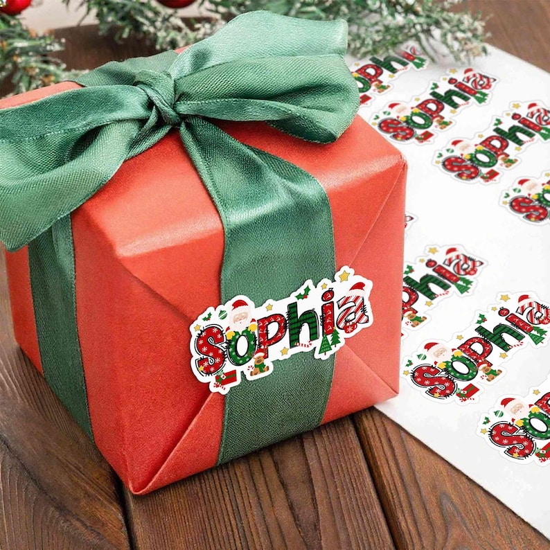 May include: A wrapped red gift box with a green ribbon and bow. A sticker with the name "Sophia" in festive colors and a Santa Claus illustration is affixed to the ribbon. Additional stickers are visible.