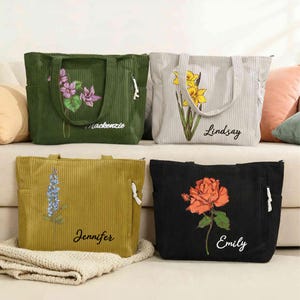 Puede incluir: Cuatro bolsos tote de pana en verde, gris, amarillo mostaza y negro. Cada bolso presenta un diseño floral y un nombre: Mackenzie, Lindsay, Jennifer y Emily. Los bolsos tienen bolsillos laterales y cremalleras blancas.
