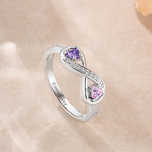 Puede incluir: Anillo plateado con diseño de símbolo de infinito. El anillo presenta dos piedras preciosas en forma de corazón, una púrpura y otra rosa, y está adornado con pequeñas piedras transparentes. Las palabras "Forever love" están grabadas en la banda.
