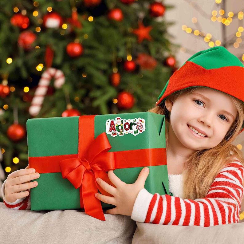 May include: A young person wearing a red and green elf hat and striped shirt holds a wrapped gift. The gift is green with a red ribbon and bow. A sticker on the gift reads "Agora" in festive lettering, with Christmas-themed illustrations.